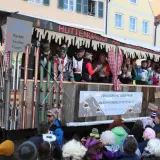 Narren feiern beim Faschingsumzug mit Skimotto. Ein Wagen im Stil einer Skihütte, dekoriert mit Skiern und Schildern wie "Parken, Saufen, Feiern", zieht durch die Gassen.