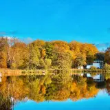 Herbstliche Farben im Schlosspark Putbus spiegeln sich im ruhigen Wasser. Ein Pavillon und ein modernes Gebäude sind zu sehen.