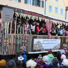Narren feiern beim Faschingsumzug mit Skimotto. Ein Wagen im Stil einer Skihütte, dekoriert mit Skiern und Schildern wie "Parken, Saufen, Feiern", zieht durch die Gassen.