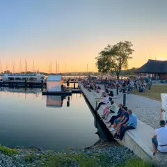 Abendstimmung beim Selliner Hafenfest. Menschen sitzen am Wasser, Boote liegen im Hafen, die Sonne geht unter.