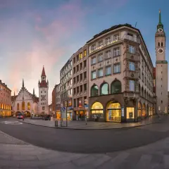 Abenddämmerung in Lehel, München. Die Deutsche Bank und das Ludwig Beck Gebäude erstrahlen im warmen Licht, während der Turm der Frauenkirche in den rosafarbenen Himmel ragt.