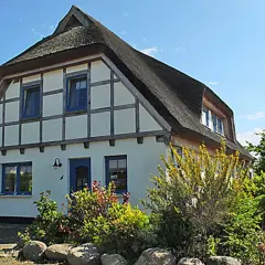Reetgedecktes Haus mit blauen Fenstern und Tür, umgeben von blühenden Sträuchern und Bäumen. Ein idyllischer Anblick am Breeger Bodden.