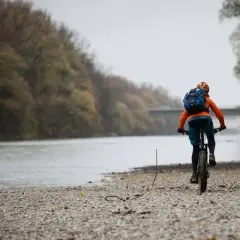 Radfahrer auf den Isar Trails, mit Rucksack und Helm, fährt am Flussufer entlang.