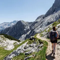 Frau mit Rucksack und Wanderstöcken genießt das Wandern in den Alpen auf einem steinigen Pfad mit Blick auf majestätische Berge.