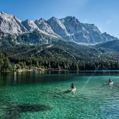 Ein Mann paddelt auf einem SUP-Board auf dem kristallklaren, blauen See in Bayern. Im Hintergrund ragen majestätische Berge auf.