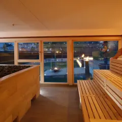Entspannter **Sauna Bereich** mit Holzbänken und Blick auf Feuerstellen im Garten. Eine Frau in einem Handtuch sitzt auf der obersten Bank.