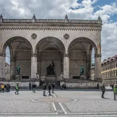 Der Odeonsplatz in München mit der Feldherrnhalle und ihren Arkaden. Menschen schlendern über den Platz, während die Sonne durch die Wolken bricht.