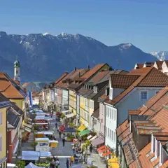 Bunte Häuserfronten säumen eine belebte Straße mit Marktständen, im Hintergrund majestätische Berge. Entdecke Murnau Geheimtipps!