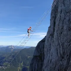 Eine Frau klettert auf einem Himmelsleiter Klettersteig eine steile Felswand empor. Die Aussicht auf die Berge ist atemberaubend.