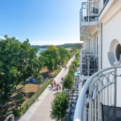 Blick von einem Balkon auf die belebte Binz Promenade mit Bäumen, Meer und Menschen, die spazieren gehen.