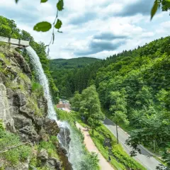 Der Trusetaler Wasserfall stürzt von einem Felsen herab. Ein Weg führt am Wasserfall vorbei, daneben eine Straße und ein bewaldeter Hang.