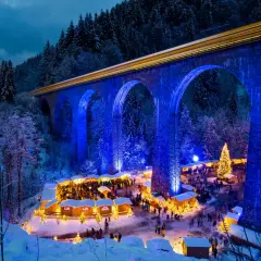 Magischer weihnachtsmarkt in deutschland unter einer beleuchteten Brücke. Ein Zug fährt vorbei, während Besucher die festliche Atmosphäre genießen.
