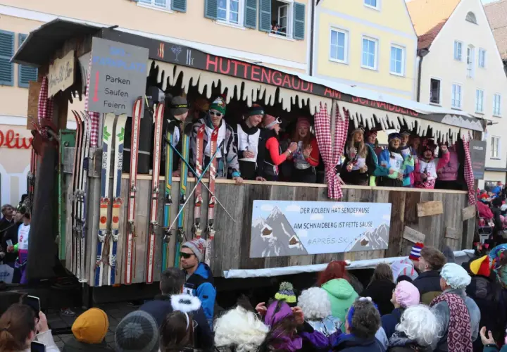Narren feiern beim Faschingsumzug mit Skimotto. Ein Wagen im Stil einer Skihütte, dekoriert mit Skiern und Schildern wie "Parken, Saufen, Feiern", zieht durch die Gassen.