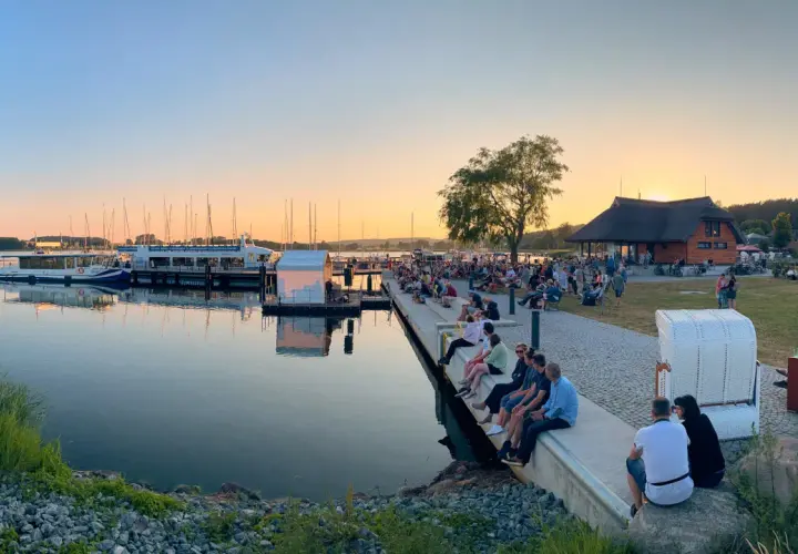 Abendstimmung beim Selliner Hafenfest. Menschen sitzen am Wasser, Boote liegen im Hafen, die Sonne geht unter.