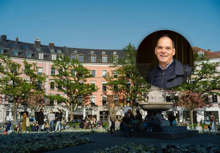Ein Mann lächelt vor einem Brunnen und rosa Gebäuden im Glockenbachviertel München.