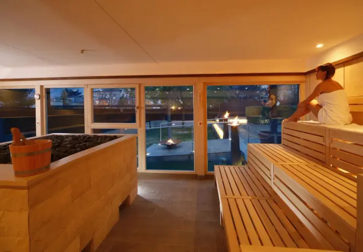 Entspannter **Sauna Bereich** mit Holzbänken und Blick auf Feuerstellen im Garten. Eine Frau in einem Handtuch sitzt auf der obersten Bank.
