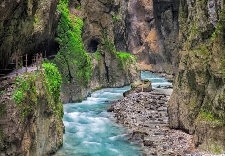 Türkisblaues Wasser rauscht durch die enge Klamm in Bayern. Ein schmaler Weg führt entlang der moosbewachsenen Felswände.