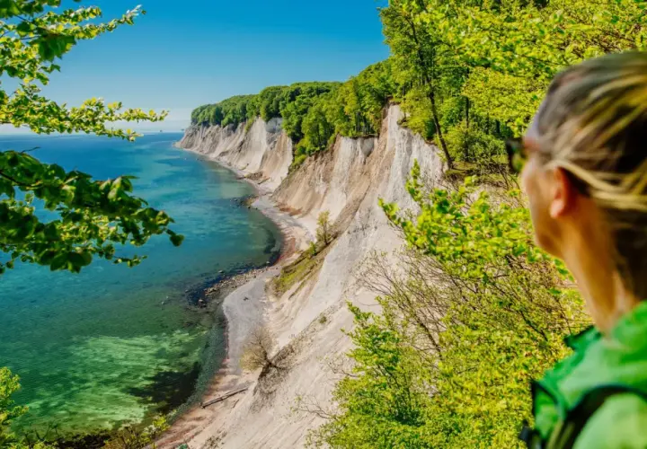 Frau blickt auf Kreidefelsen und das Meer bei Reddevitz. Grüne Bäume umrahmen die malerische Küstenlandschaft.