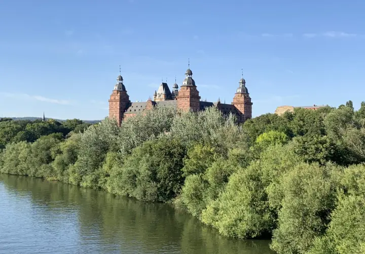 Schloss Johannisberg, ein malerisches Ziel im Spessart, ragt über üppige Bäume am Flussufer auf.