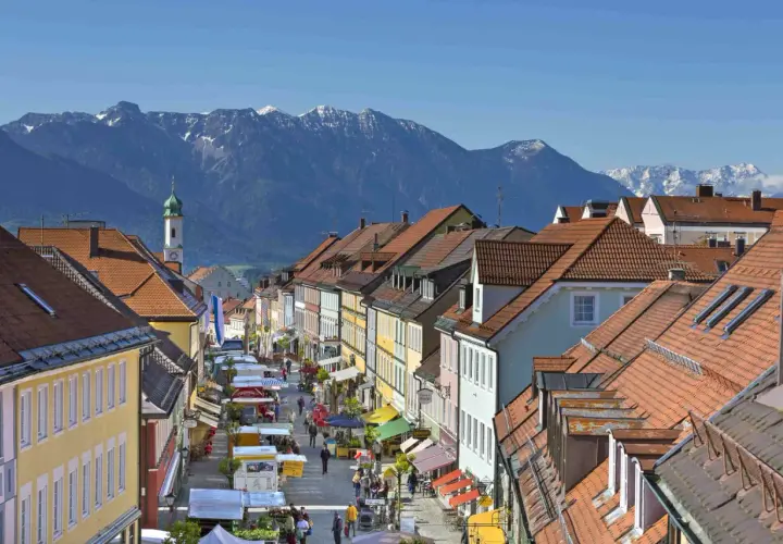 Bunte Häuserfronten und ein Markt beleben die Gasse in Murnau am Staffelsee. Im Hintergrund ragen schneebedeckte Berge auf.