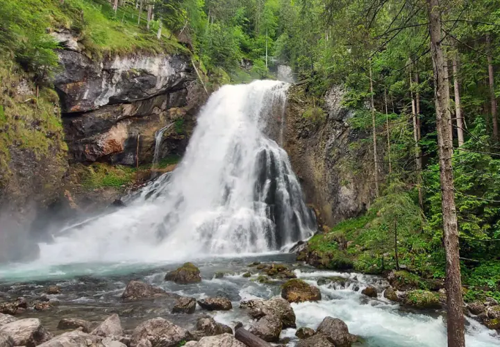 Tosender Wasserfall im Teutoburger Wald, umgeben von Felsen und üppigem Grün. Das Wasser stürzt tosend in ein türkisfarbenes Becken.