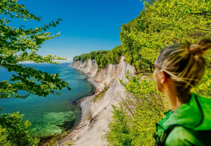 Frau blickt auf Kreidefelsen im Nationalpark Jasmund. Türkisfarbenes Meer und grüne Bäume.