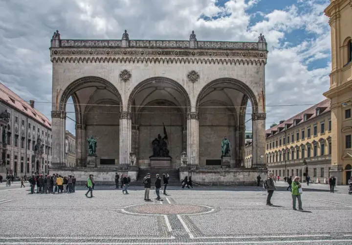 Der Odeonsplatz in München mit der Feldherrnhalle und ihren Arkaden. Menschen schlendern über den Platz, während die Sonne durch die Wolken bricht.