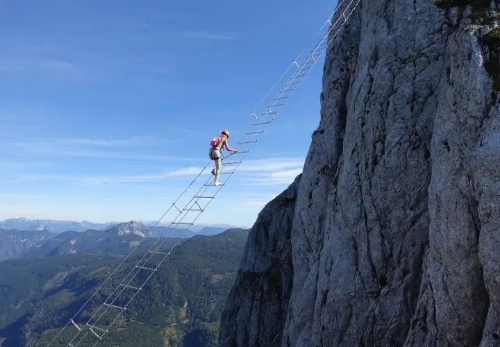 Eine Frau klettert auf einem Himmelsleiter Klettersteig eine steile Felswand empor. Die Aussicht auf die Berge ist atemberaubend.