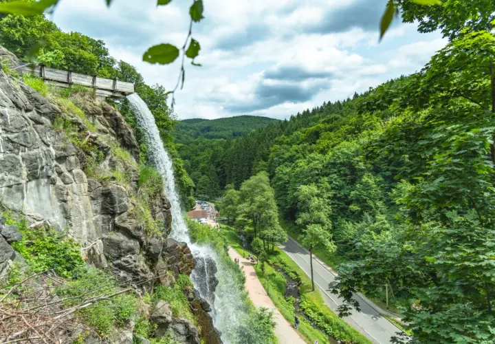 Der Trusetaler Wasserfall stürzt von einem Felsen herab. Ein Weg führt am Wasserfall vorbei, daneben eine Straße und ein bewaldeter Hang.