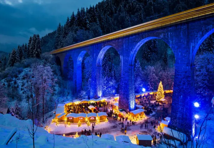 Magischer weihnachtsmarkt in deutschland unter einer beleuchteten Brücke. Ein Zug fährt vorbei, während Besucher die festliche Atmosphäre genießen.