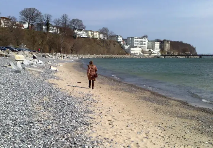 Eine Frau spaziert am Strand entlang, im Hintergrund die Bäderarchitektur von Sassnitz.