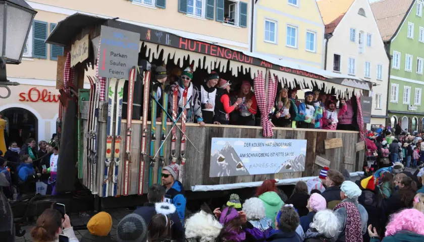 Narren feiern beim Faschingsumzug mit Skimotto. Ein Wagen im Stil einer Skihütte, dekoriert mit Skiern und Schildern wie "Parken, Saufen, Feiern", zieht durch die Gassen.