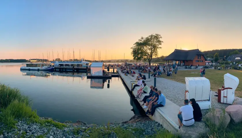 Abendstimmung beim Selliner Hafenfest. Menschen sitzen am Wasser, Boote liegen im Hafen, die Sonne geht unter.