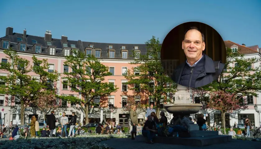 Ein Mann lächelt vor einem Brunnen und rosa Gebäuden im Glockenbachviertel München.
