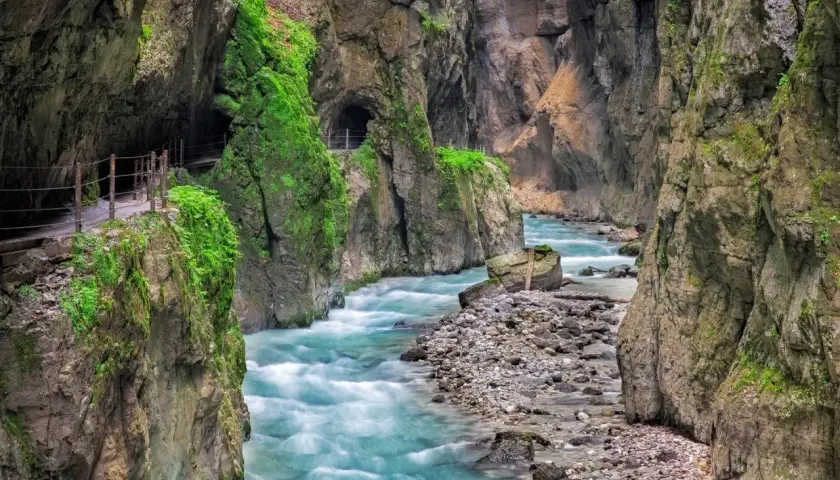 Türkisblaues Wasser rauscht durch die enge Klamm in Bayern. Ein schmaler Weg führt entlang der moosbewachsenen Felswände.