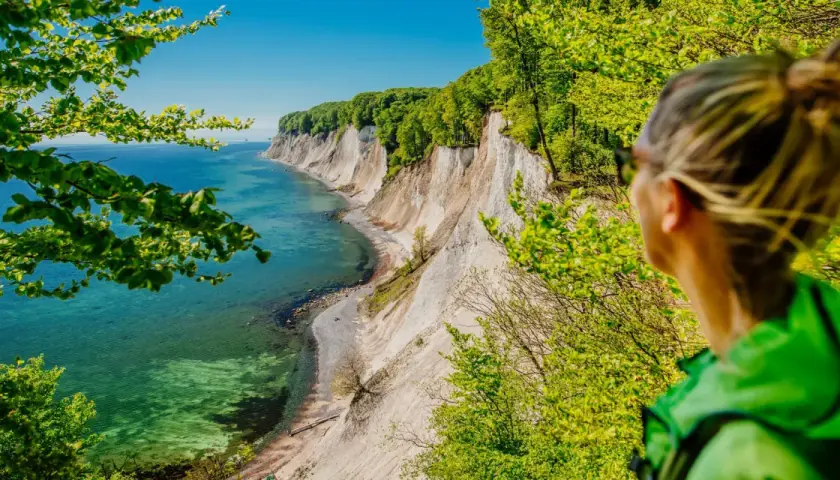 Frau blickt auf Kreidefelsen und das Meer bei Reddevitz. Grüne Bäume umrahmen die malerische Küstenlandschaft.