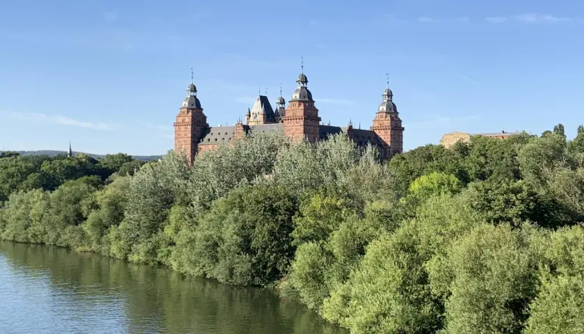 Schloss Johannisberg, ein malerisches Ziel im Spessart, ragt über üppige Bäume am Flussufer auf.