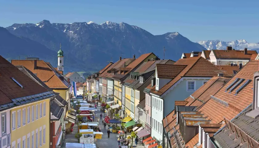 Bunte Häuserfronten und ein Markt beleben die Gasse in Murnau am Staffelsee. Im Hintergrund ragen schneebedeckte Berge auf.