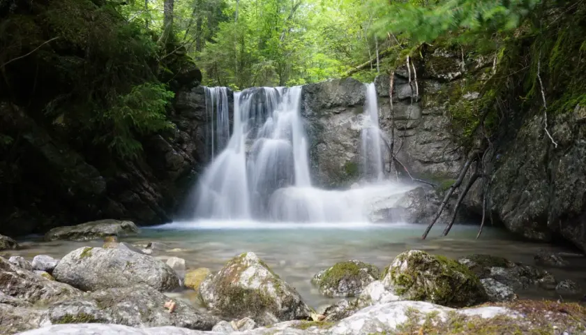 Ein malerischer oberammergau wasserfall stürzt über moosbewachsene Felsen in ein klares Becken, umgeben von üppigem Grün.
