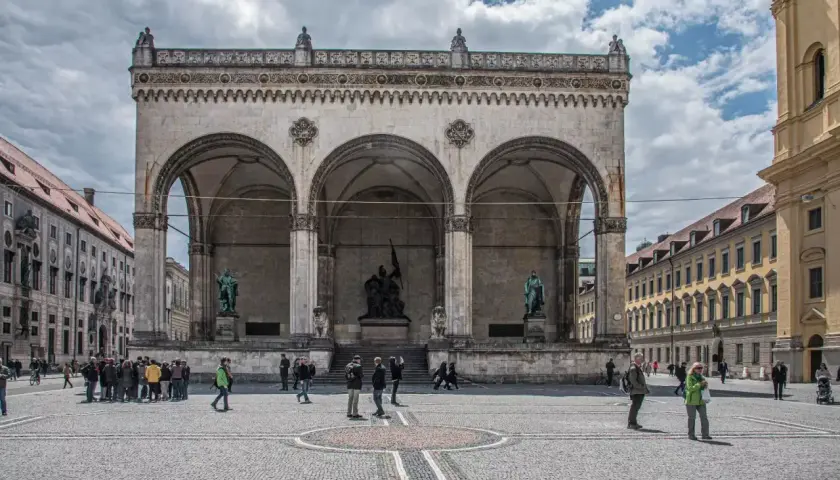 Der Odeonsplatz in München mit der Feldherrnhalle und ihren Arkaden. Menschen schlendern über den Platz, während die Sonne durch die Wolken bricht.