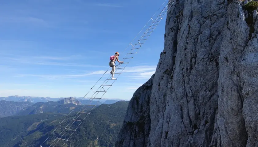 Eine Frau klettert auf einem Himmelsleiter Klettersteig eine steile Felswand empor. Die Aussicht auf die Berge ist atemberaubend.