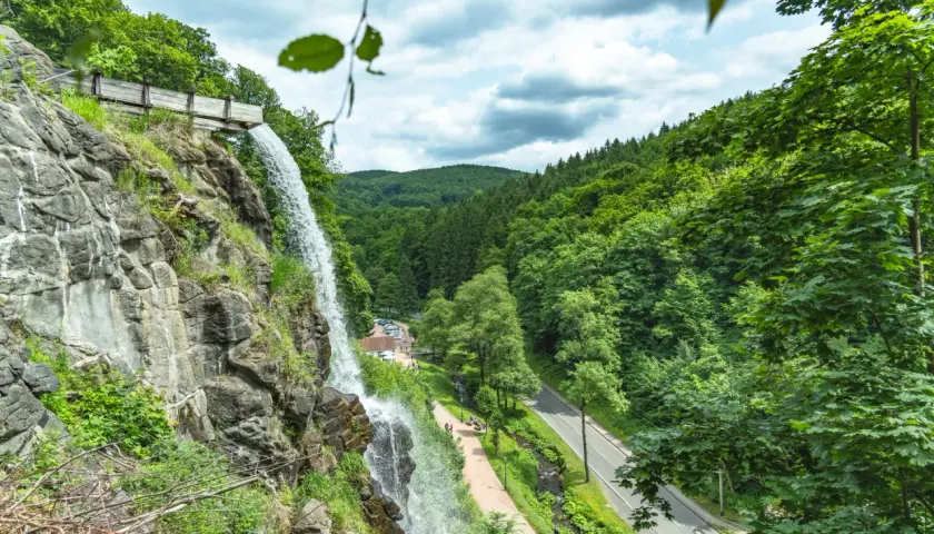 Der Trusetaler Wasserfall stürzt von einem Felsen herab. Ein Weg führt am Wasserfall vorbei, daneben eine Straße und ein bewaldeter Hang.