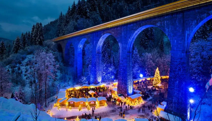 Magischer weihnachtsmarkt in deutschland unter einer beleuchteten Brücke. Ein Zug fährt vorbei, während Besucher die festliche Atmosphäre genießen.
