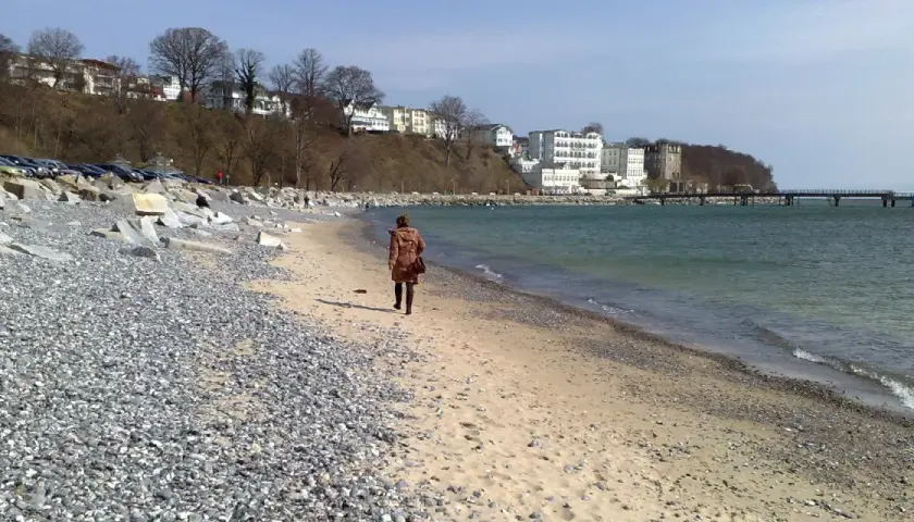 Eine Frau spaziert am Strand entlang, im Hintergrund die Bäderarchitektur von Sassnitz.