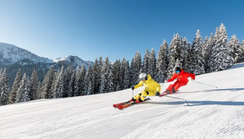 Zwei Skifahrer in leuchtenden Farben sausen über eine präparierte Piste in den **Skigebieten Alpen**. Schneebedeckte Tannen und Berge im Hintergrund.