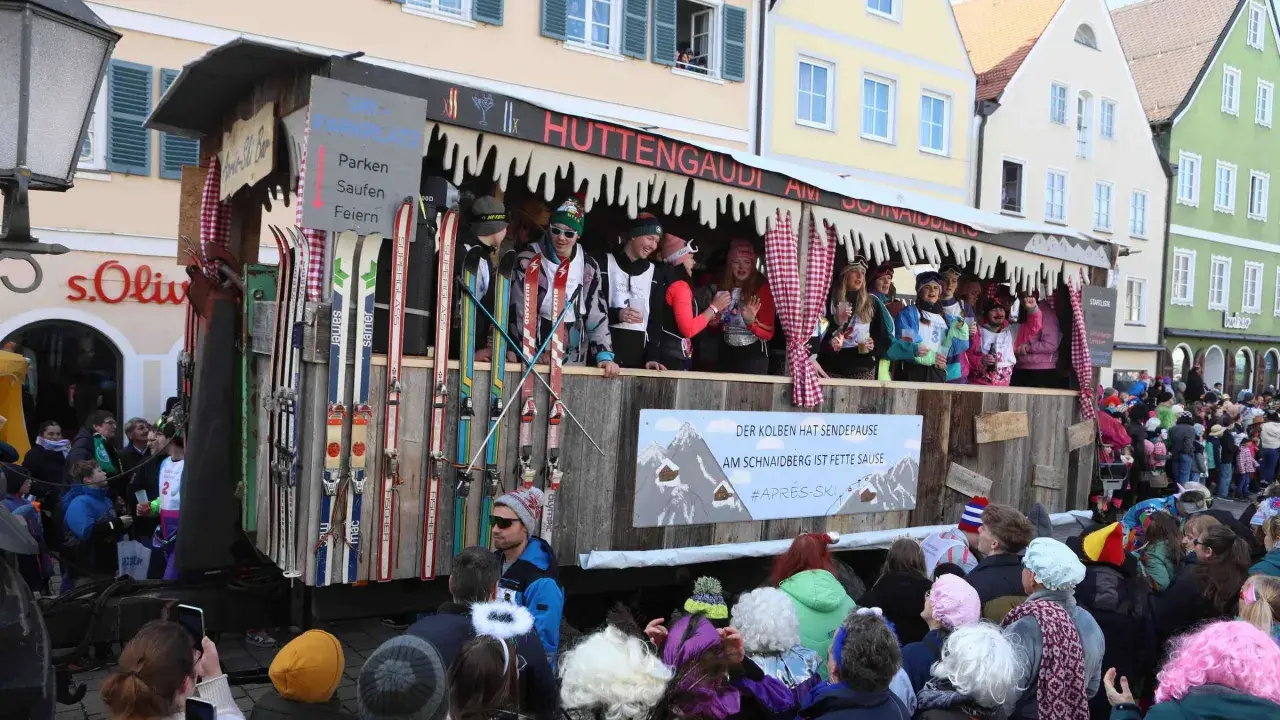 Narren feiern beim Faschingsumzug mit Skimotto. Ein Wagen im Stil einer Skihütte, dekoriert mit Skiern und Schildern wie "Parken, Saufen, Feiern", zieht durch die Gassen.