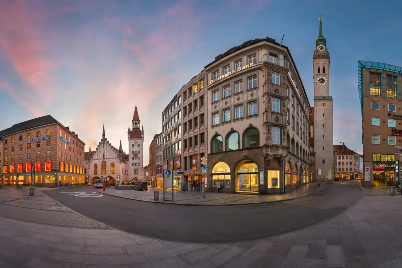 Abenddämmerung in Lehel, München. Die Deutsche Bank und das Ludwig Beck Gebäude erstrahlen im warmen Licht, während der Turm der Frauenkirche in den rosafarbenen Himmel ragt.