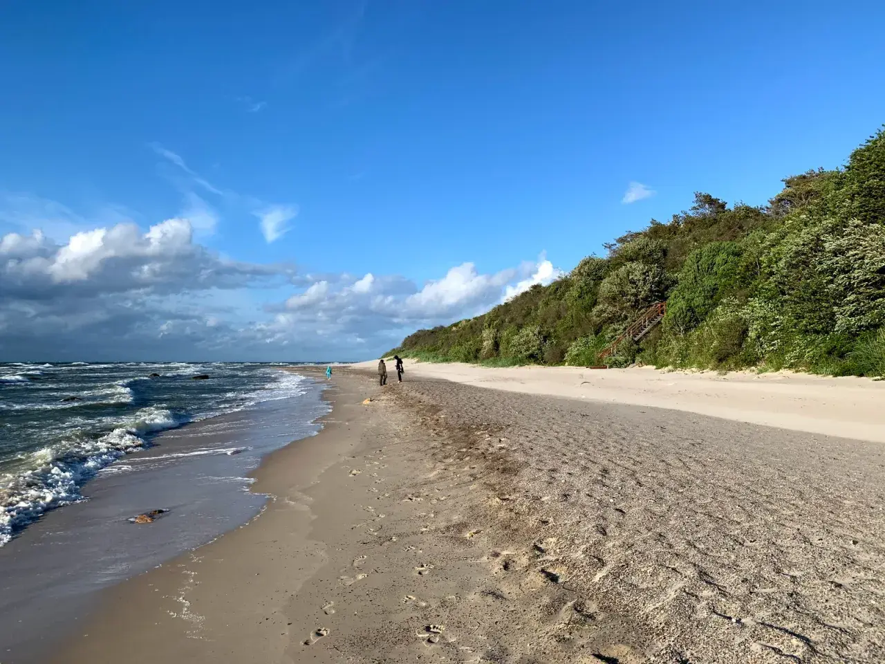 Spaziergang am Strand von Dranske Bakenberg. Wellen brechen an der Küste, während zwei Personen den Sandweg entlanggehen.