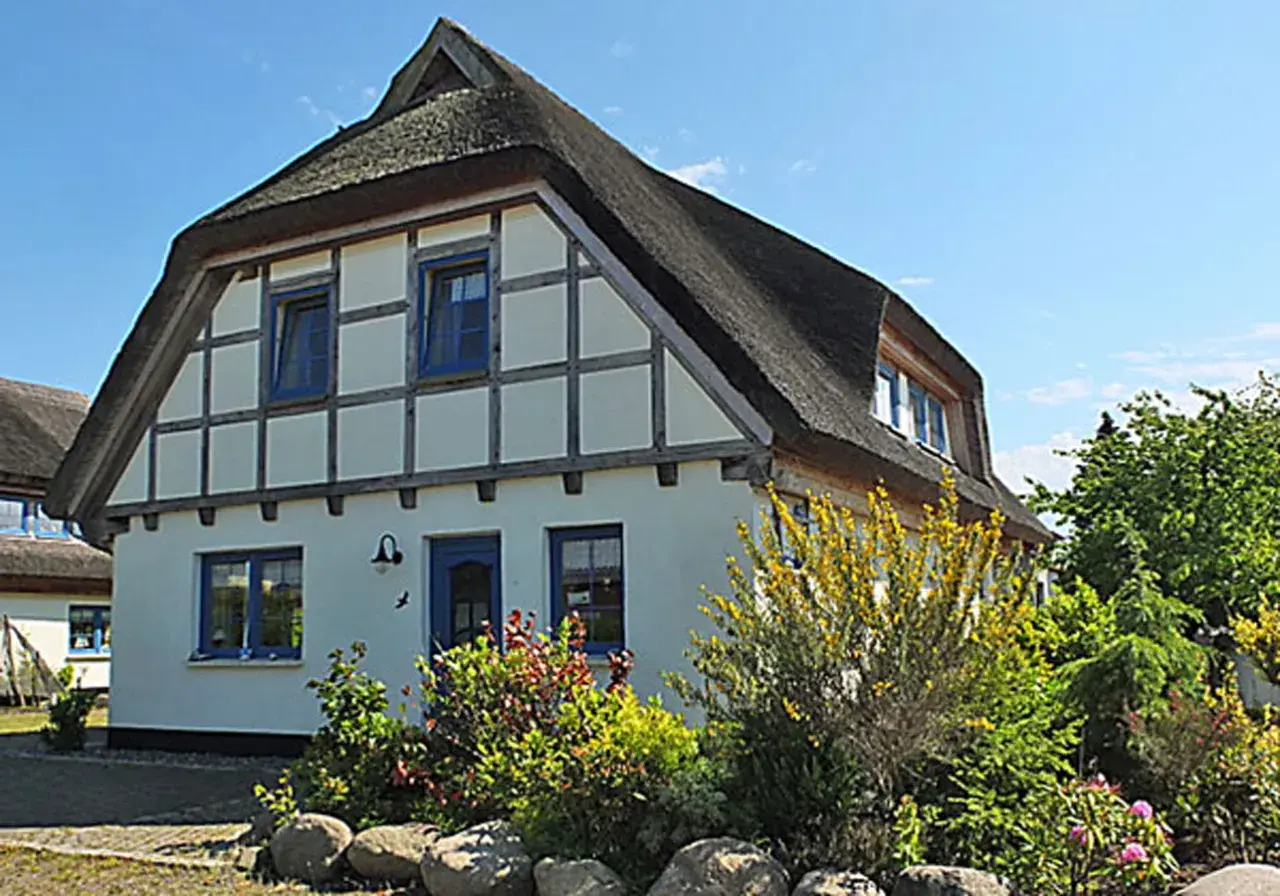 Reetgedecktes Haus mit blauen Fenstern und Tür, umgeben von blühenden Sträuchern und Bäumen. Ein idyllischer Anblick am Breeger Bodden.