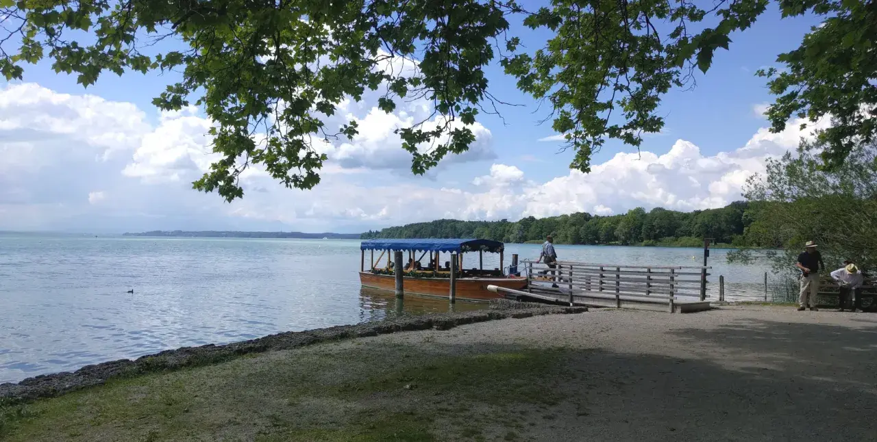Ein Ausflugsboot wartet am Steg am Starnberger See. Zwei Männer sitzen auf einer Bank im Schatten.